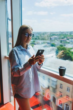 Modern kadın, büyük bir pencerede durup kahve içerken telefonla konuşuyor. Modern başarılı kadın kavramı. Açık bir ofiste genç bir kız