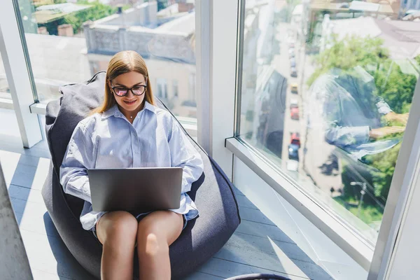 Young millennial woman in classic eye protection glasses connecting to WIFI internet on laptop in coworking space to browse web information on digital netbooks, european woman chatting