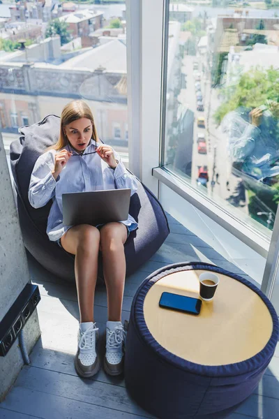 A modern woman watches videos on a laptop and drinks coffee. The concept of distance or e-learning. A young smiling woman sits in a chair at the table.