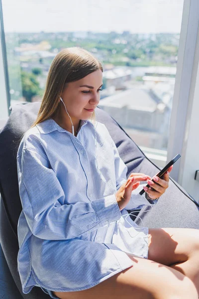 Woman in classic glasses listening to music with headphones from phone and smiling while sitting in modern coworking space, carefree millennial woman in glasses enjoying leisure time for communication