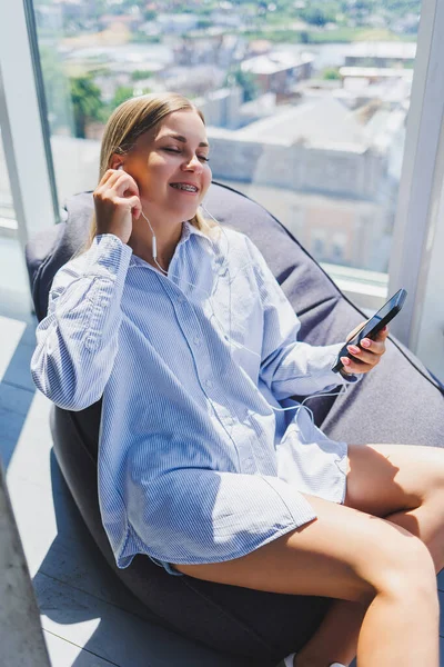 Woman in classic glasses listening to music with headphones from phone and smiling while sitting in modern coworking space, carefree millennial woman in glasses enjoying leisure time for communication