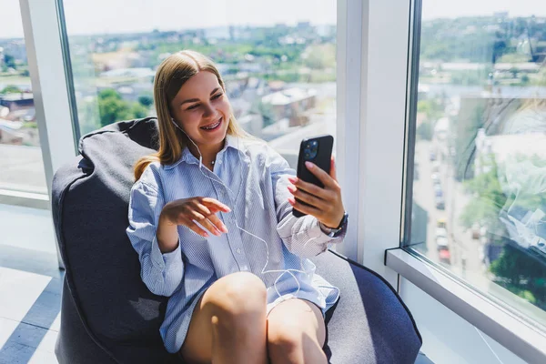 Woman in classic glasses listening to music with headphones from phone and smiling while sitting in modern coworking space, carefree millennial woman in glasses enjoying leisure time for communication