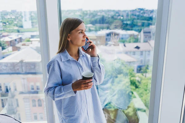 Modern woman freelancer talking on the phone while standing at a large window and drinking coffee. The concept of a modern successful woman. Young girl in an open office