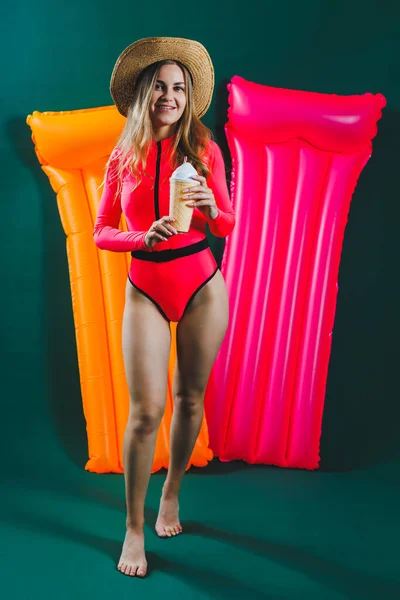 A young woman in a bright pink swimsuit and hat with an inflatable pink mattress on a green background. Summer rest. Girl with a cocktail in her hands in a bathing suit