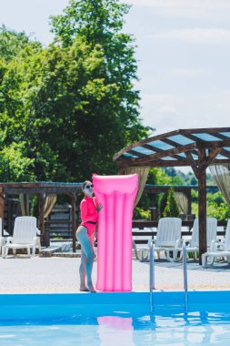 Pembe mayolu ve şişme yataklı güzel bir kadın büyük bir havuzun yanında duruyor. Yaz tatillerinde yüzme havuzlu bir otelde. Mayolu ve güneş gözlüklü genç kız.