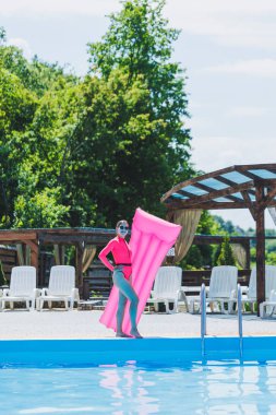 Pembe mayolu ve şişme yataklı güzel bir kadın büyük bir havuzun yanında duruyor. Yaz tatillerinde yüzme havuzlu bir otelde. Mayolu ve güneş gözlüklü genç kız.