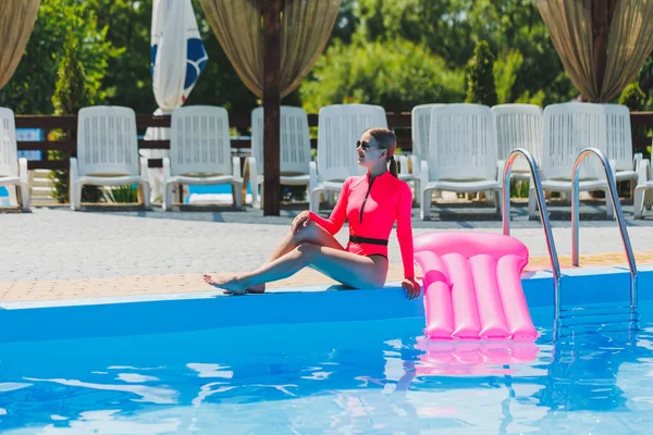 Pembe mayolu ve şişme yataklı güzel bir kadın büyük bir havuzun yanında duruyor. Yaz tatillerinde yüzme havuzlu bir otelde. Mayolu ve güneş gözlüklü genç kız.