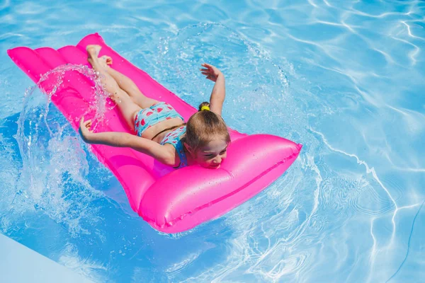 7-8 yaşlarında bir kız şişme pembe bir yatakta havuzda yüzüyor. Yaz tatili