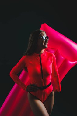 Pembe mayolu çekici genç bir kadın, koyu bir arka planda izole edilmiş. Tatil konsepti, yaz tatili. Güneş gözlüklü ince bir kız elinde şişme pembe bir yatak tutuyor.