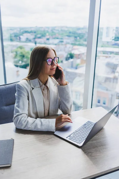 A woman manager in glasses sits at a laptop in an office with a stunning view of the city and conducts a business conversation on the phone. Business lady working at the wooden table in the office