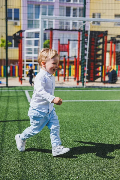 The child runs in the park. A boy in a white shirt and trousers. Green lawn or field. Mowed lawn. Walk and play in the forest. Active sports.
