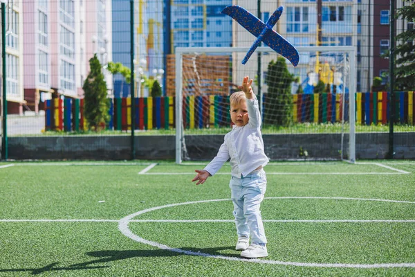 A child runs in the park with a toy airplane. A boy in a white shirt and trousers. Green lawn or field. Mowed lawn. Walk and play in the forest. Active sports.