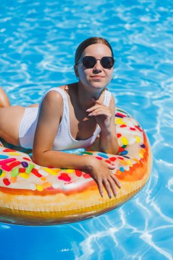 Havuzda dinlen. Mayolu, güneş gözlüklü ve mavi suda yüzen şişme lastik yüzüklü mutlu genç bir kadın. Spa tatil merkezinde yaz tatili