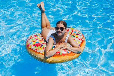 Havuzda dinlen. Mayolu, güneş gözlüklü ve mavi suda yüzen şişme lastik yüzüklü mutlu genç bir kadın. Spa tatil merkezinde yaz tatili