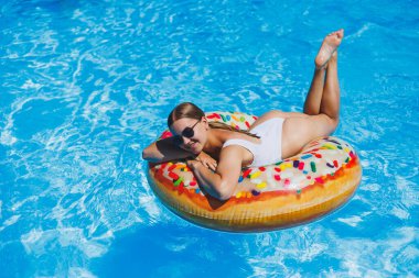 Beyaz mayo ve güneş gözlüğü takmış ince bir kadın şişme dairenin içinde havuzda yatıyor. Yaz tatili.