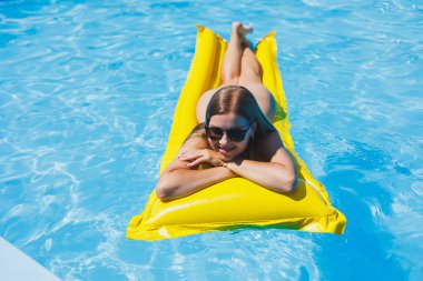 Beautiful young woman in sunglasses on a yellow inflatable mattress in a swimsuit swims in the pool of a luxury hotel, summer vacation, travel, smile