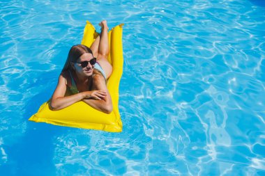 Güneş gözlüklü, sarı şişme yatakta mayo giymiş güzel bir kadın lüks bir otelin havuzunda yüzüyor, yaz tatili, seyahat, gülümseme.