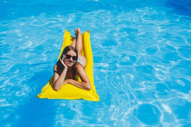 Beautiful young woman in sunglasses on a yellow inflatable mattress in a swimsuit swims in the pool of a luxury hotel, summer vacation, travel, smile