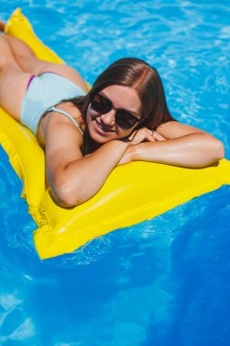 Summer lifestyle portrait of beautiful woman in sunglasses floating on inflatable mattress in outdoor swimming pool. Summer vacation on a tropical island.