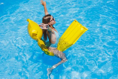 Beautiful young woman in sunglasses on a yellow inflatable mattress in a swimsuit swims in the pool of a luxury hotel, summer vacation, travel, smile