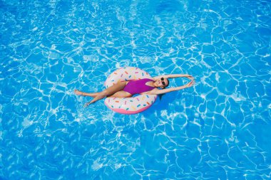 Su parkında yüzen mayo giymiş, parlak mavi bir havuzda yüzen ve kameraya gülümseyen güzel, sıska genç bir kadın. Yaz tatili.