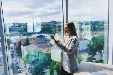 Attractive female blogger in a jacket with a modern smartphone in her hands, thoughtful and daydreaming, a young blogger holding a mobile phone, reflects on social media posts