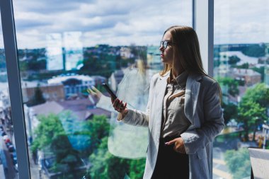 Elinde modern bir akıllı telefon olan ceketli çekici bir kadın blogcu, düşünceli ve hayal kuran, cep telefonu tutan genç bir blogcu, sosyal medya paylaşımlarına yansıyor.