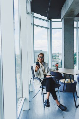 Elinde modern bir akıllı telefon olan ceketli çekici bir kadın blogcu, düşünceli ve hayal kuran, cep telefonu tutan genç bir blogcu, sosyal medya paylaşımlarına yansıyor.