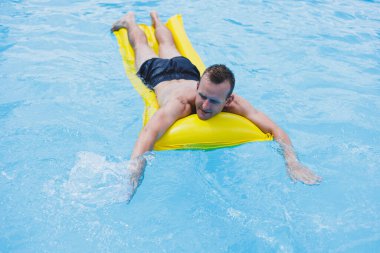 A young man in shorts enjoys the water park floating in an inflatable big ring on a sparkling blue pool smiling at the camera. Summer vacation.