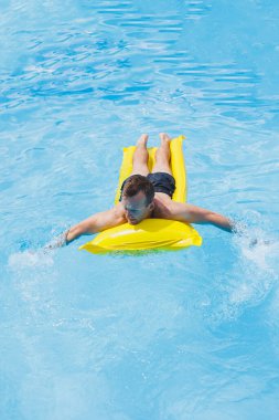 Şortlu genç bir adam, kameraya gülümseyen parlak mavi bir havuzda şişme bir yüzükle su parkının keyfini çıkarıyor. Yaz tatili.