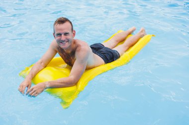 Şortlu genç bir adam, kameraya gülümseyen parlak mavi bir havuzda şişme bir yüzükle su parkının keyfini çıkarıyor. Yaz tatili.