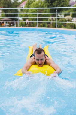 A young man in shorts enjoys the water park floating in an inflatable big ring on a sparkling blue pool smiling at the camera. Summer vacation.