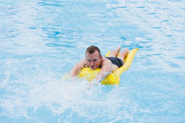 A young man in shorts enjoys the water park floating in an inflatable big ring on a sparkling blue pool smiling at the camera. Summer vacation.