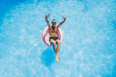 Güneş gözlüklü ve şortlu genç bir adam havuzda şişme bir çöreğin üzerinde dinleniyor. Yaz tatili