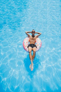 Güneş gözlüklü ve şortlu genç bir adam havuzda şişme bir çöreğin üzerinde dinleniyor. Yaz tatili