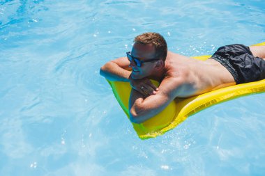 Attractive young man in sunglasses and shorts is relaxing on an inflatable yellow mattress in the pool. Summer vacation