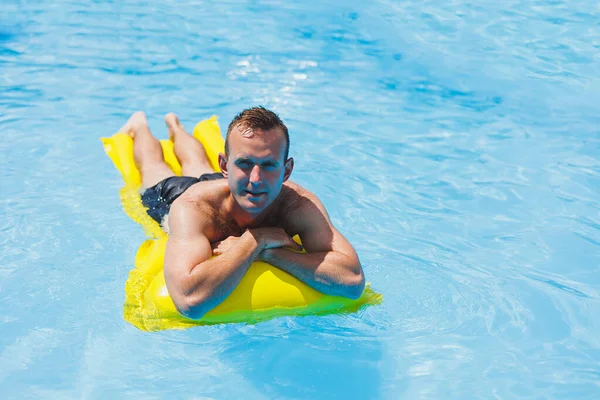 A young man in shorts enjoys the water park floating in an inflatable big ring on a sparkling blue pool smiling at the camera. Summer vacation.