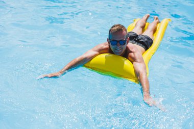 Attractive young man in sunglasses and shorts is relaxing on an inflatable yellow mattress in the pool. Summer vacation