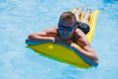 Attractive young man in sunglasses and shorts is relaxing on an inflatable yellow mattress in the pool. Summer vacation