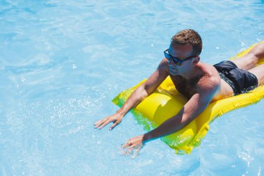 Attractive young man in sunglasses and shorts is relaxing on an inflatable yellow mattress in the pool. Summer vacation