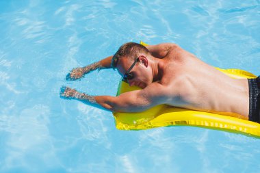 Attractive young man in sunglasses and shorts is relaxing on an inflatable yellow mattress in the pool. Summer vacation