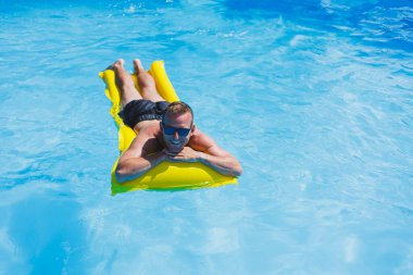Attractive young man in sunglasses and shorts is relaxing on an inflatable yellow mattress in the pool. Summer vacation