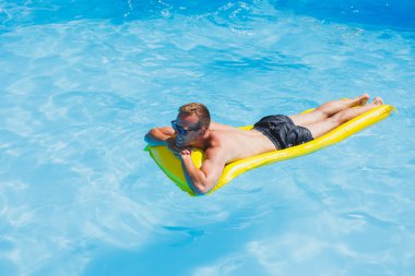 Attractive young man in sunglasses and shorts is relaxing on an inflatable yellow mattress in the pool. Summer vacation