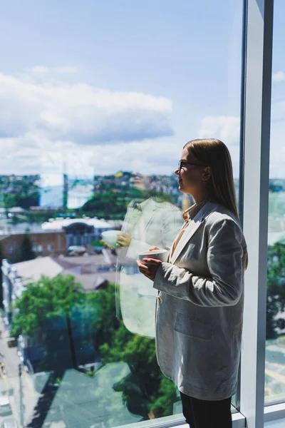 Woman manager in a jacket and glasses on her face drinking coffee during a break at work standing with a cup in the office, pensive female project planning freelancer doing remote work