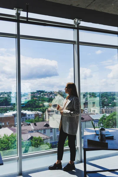 Ceketli, gözlüklü, kahve içen, kahve içen bir kadın. Ofiste elinde bir fincanla dikiliyor. Uzaktan kumandalı iş planlayan düşünceli bir kadın.