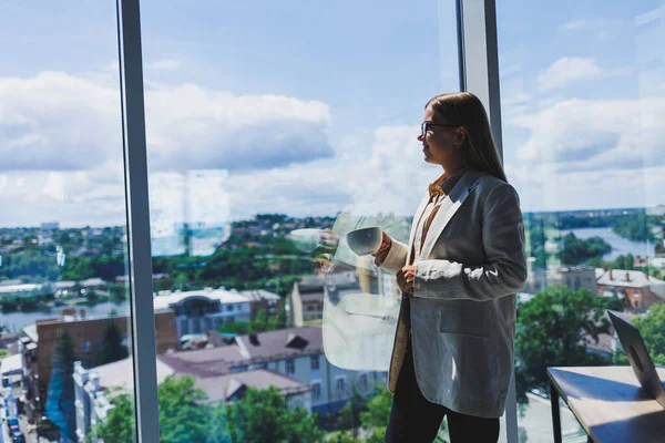 Woman manager in a jacket and glasses on her face drinking coffee during a break at work standing with a cup in the office, pensive female project planning freelancer doing remote work