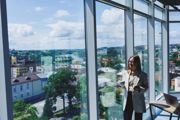 Woman manager in a jacket and glasses on her face drinking coffee during a break at work standing with a cup in the office, pensive female project planning freelancer doing remote work