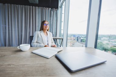 Deftere bakan güzel bir iş kadını, işine odaklanmış, serbest çalışan, uzaktan çalışan, öğrenim gören, modern ofis, kapalı alan sosyal mesafesi. Gözlüklü ve dizüstü bilgisayarı olan kadın.