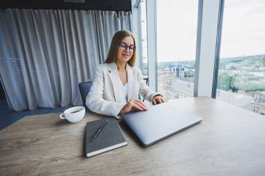 Zarif bir ceket ve optik gözlüklü başarılı genç bayan yönetici, iş yerindeki ofiste oturuyor, modern bir dizüstü bilgisayarla deneyimli bir kadın, ofiste çalışıyor.