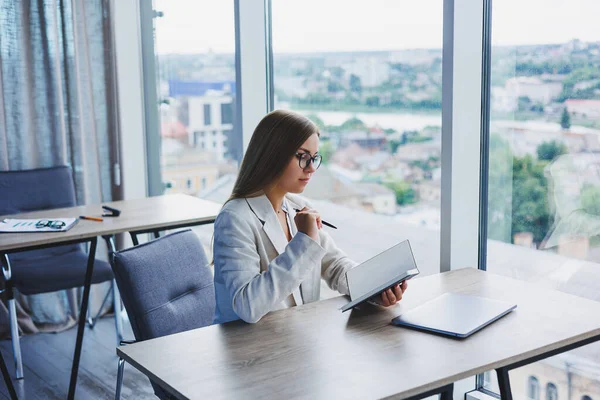 Pretty business woman looking through notebook, focused on work, freelancer, remote work, studying study, modern office, indoor social distancing. Woman in office with glasses and laptop